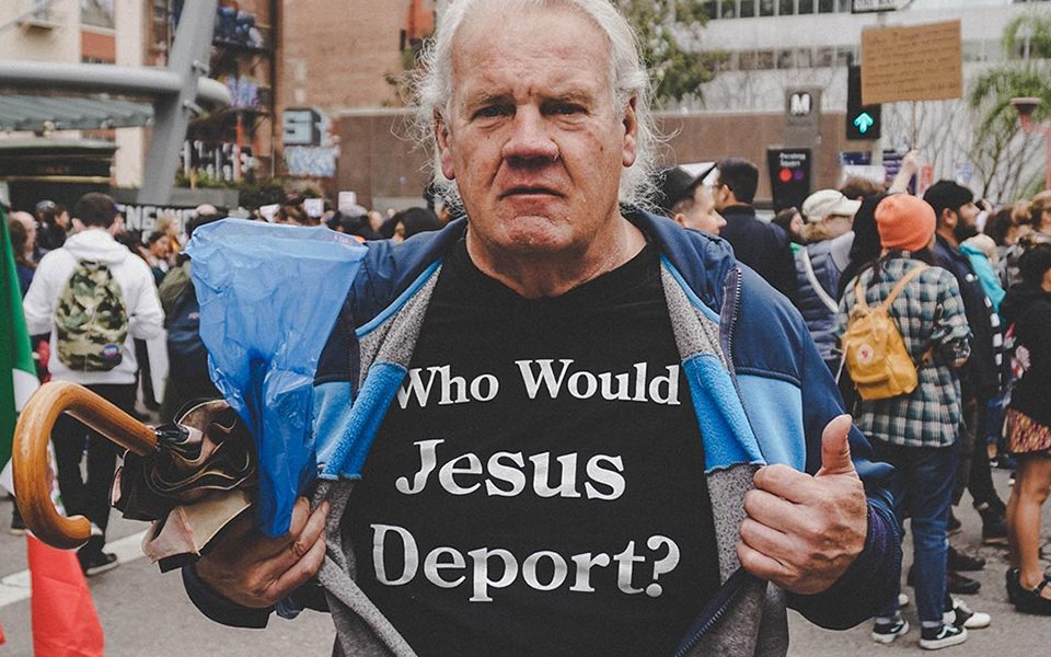 A man at a protest wearing a shirt with the text "who would jesus deport?" while holding a megaphone.
