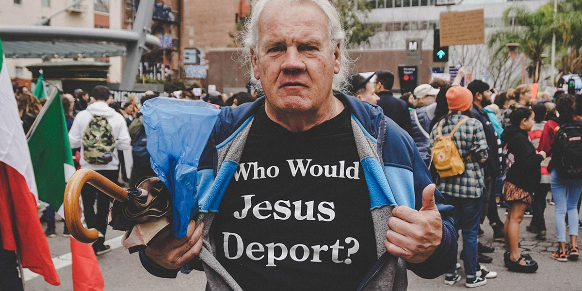 A man at a protest wearing a shirt with the text "who would jesus deport?" while holding a megaphone.