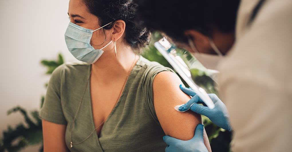 A healthcare worker administers a vaccine to a smiling woman wearing a face mask.