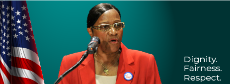 A woman in a red jacket speaking at a podium with the united states flag beside her, with the words "dignity. fairness. respect.