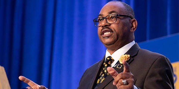 A man in glasses wearing a suit with a badge gives a speech.