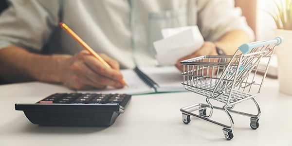 Person calculating expenses or budget with a shopping cart and calculator on a desk, symbolizing budgeting for shopping or financial planning.