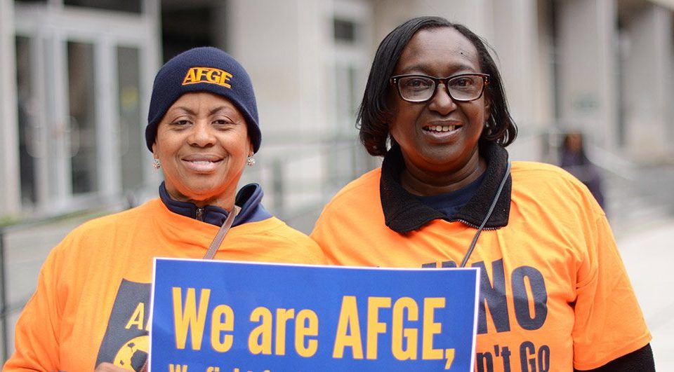 Two individuals wearing afge hats and orange shirts, one holding a sign that reads "we are afge.